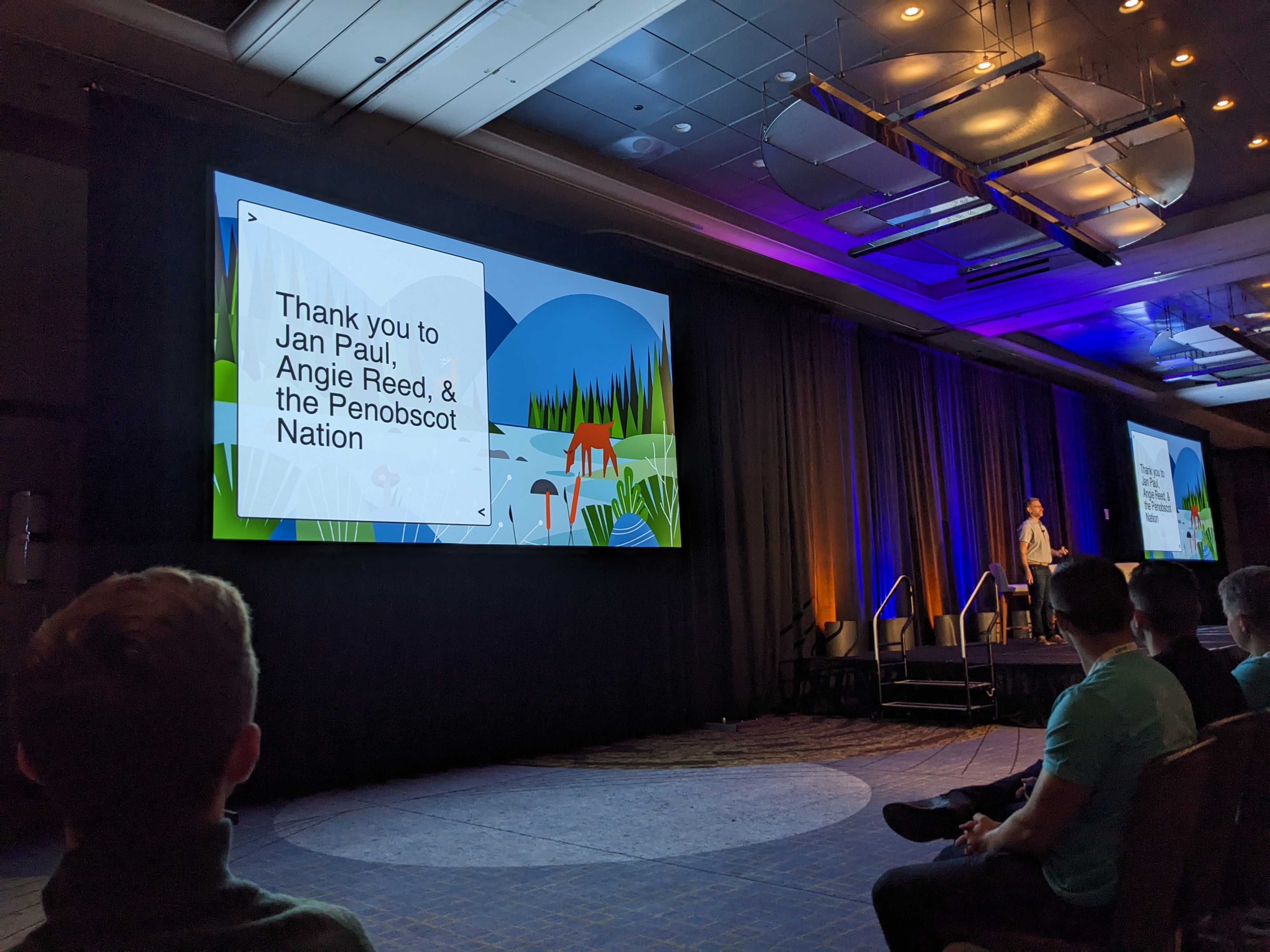 photo of posit::conf stage with screen projecting image with text "Thank you to Jan Paul, Angie Reed & the Penobscot Indian Nation" over a stylized illustration of a deer drinking from a river with green shorts and blue hills in the distance, speaker Hadley Wickham standing to right in the distance